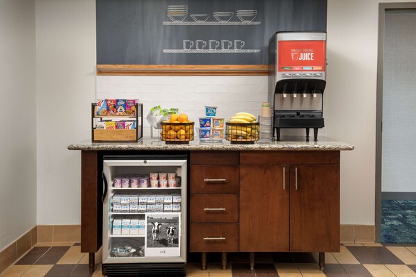 A hotel room with a kitchenette area featuring a refrigerator stocked with beverages, a coffee machine, and a fruit bowl.