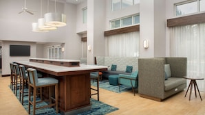 Une chambre d’hôtel moderne dotée d’un grand bar en bois, d’un coin salon avec des canapés et des fauteuils, d’un téléviseur à écran plat et de grandes fenêtres avec des rideaux.