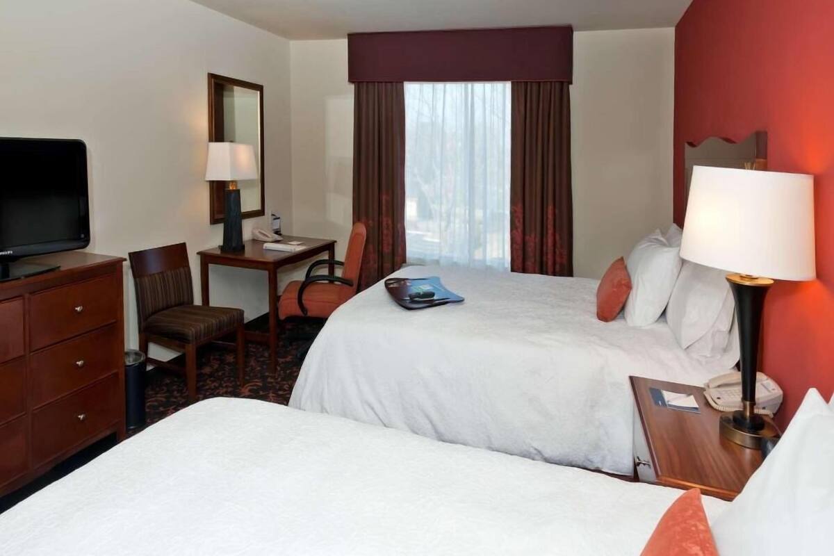 A hotel room with two beds, a desk, a chair, a television, and a window with curtains.