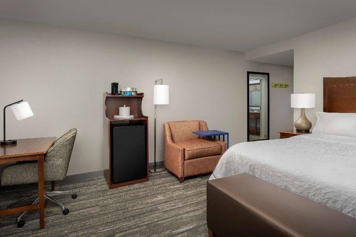 A hotel room with a bed, desk, chair, small table, and a minibar.