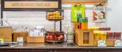 A breakfast bar with a sign reading "HAPPY BREAKFAST STARTS HERE", a fruit basket, juice dispensers, and a menu board.