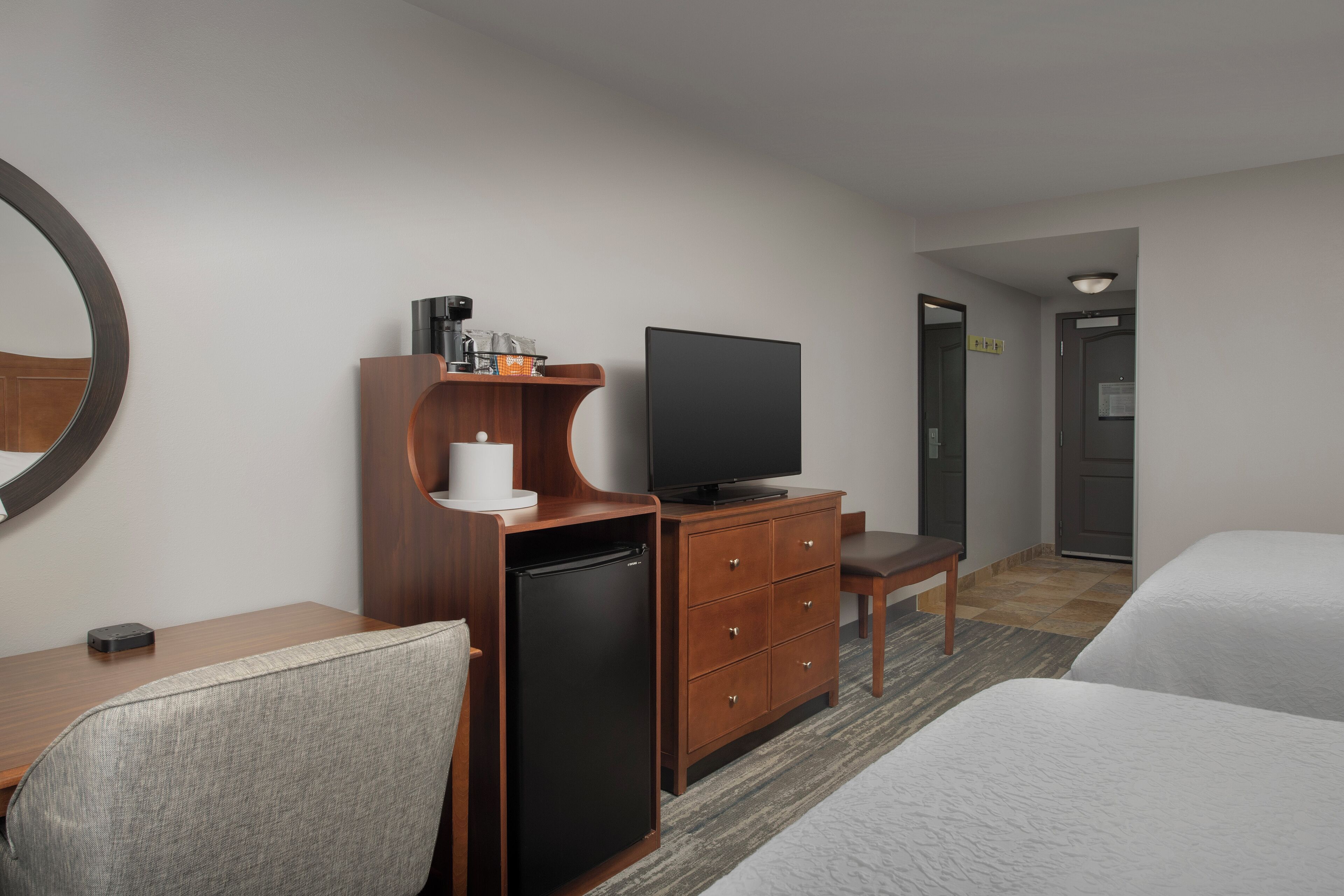 Una habitación de hotel con cama, escritorio, computadora, televisor, refrigerador pequeño y espejo.