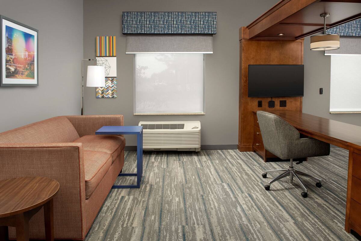 A hotel room with a sofa, a desk, a chair, a flat-screen TV, a large window with blinds, an air conditioner, and a patterned carpet.