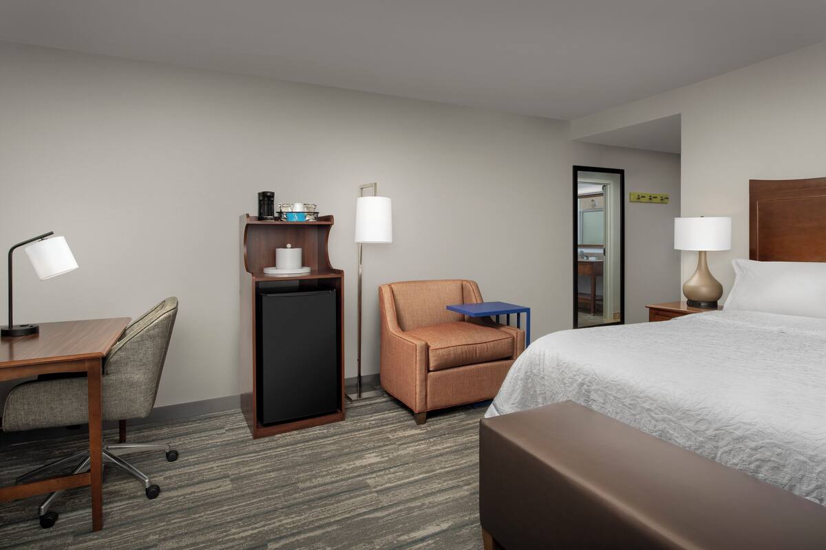 A hotel room with a bed, desk, chair, small table, lamp, and a minibar.