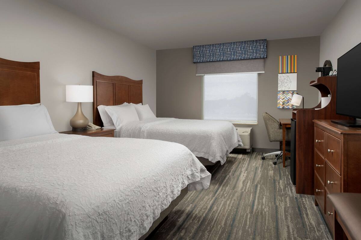 A hotel room with two beds, a desk, a chair, a television, and artwork on the walls.