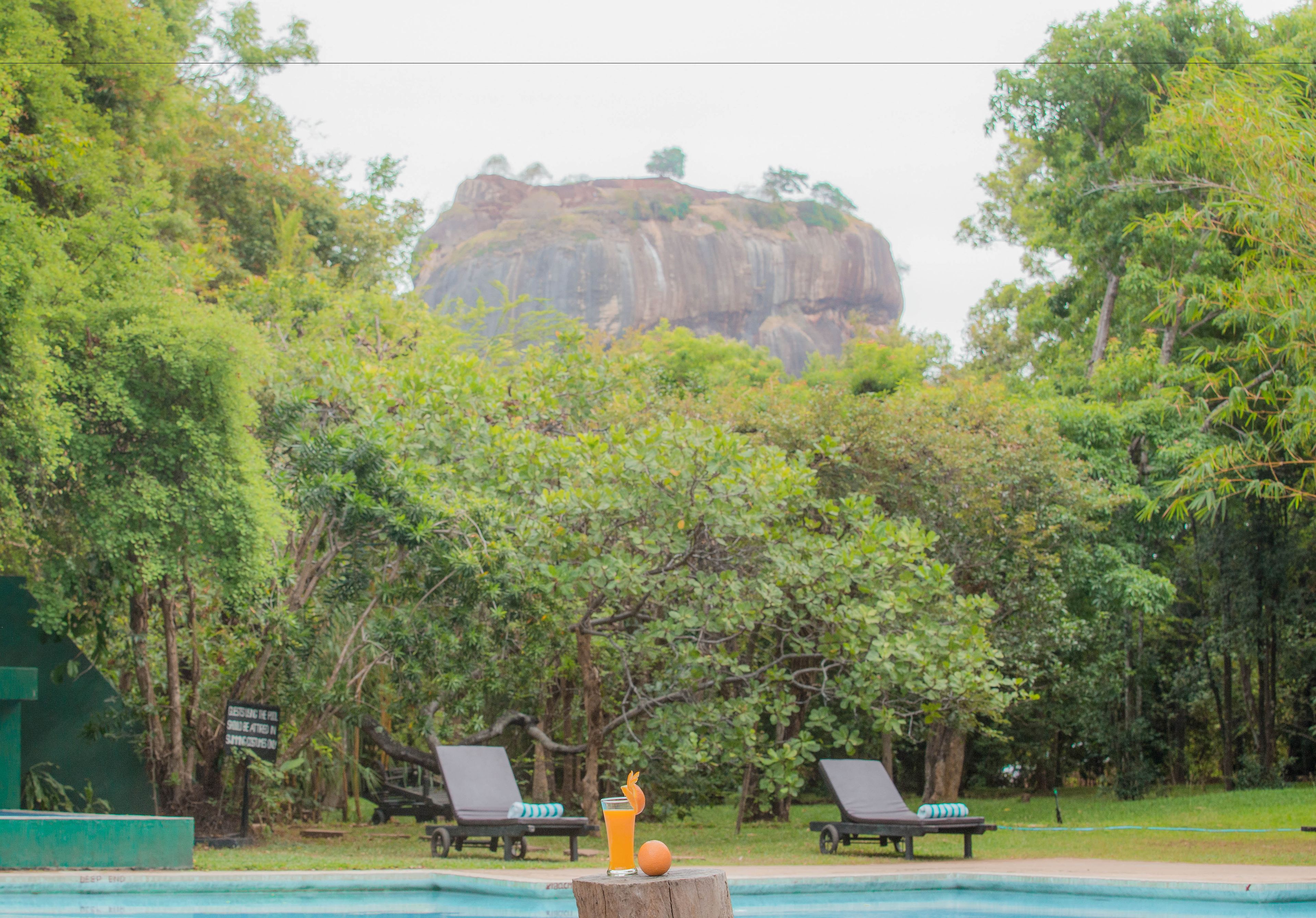 A pool area with lounge chairs and a signboard, surrounded by lush greenery and a prominent rock formation in the background.