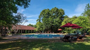 A swimming pool with lounge chairs and umbrellas, surrounded by trees and a building with a red roof. - Sigiriya Village (Sigiriya)