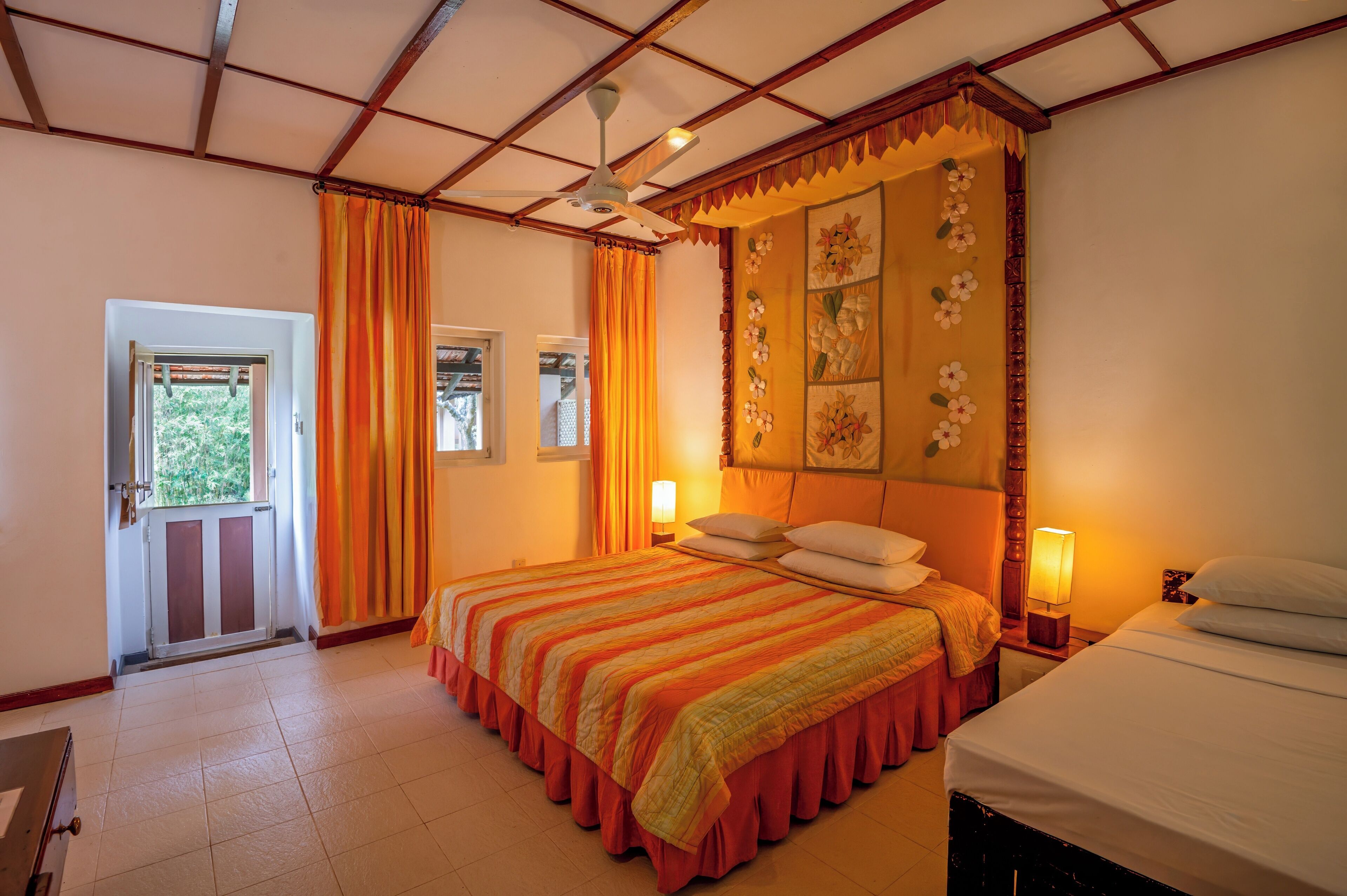 A hotel room with a large bed, two bedside tables with lamps, a ceiling fan, and a door leading to an outdoor area.