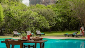 A poolside table with a drink, a basket of bread, and a plate of food, surrounded by lounge chairs and a lush green backdrop.