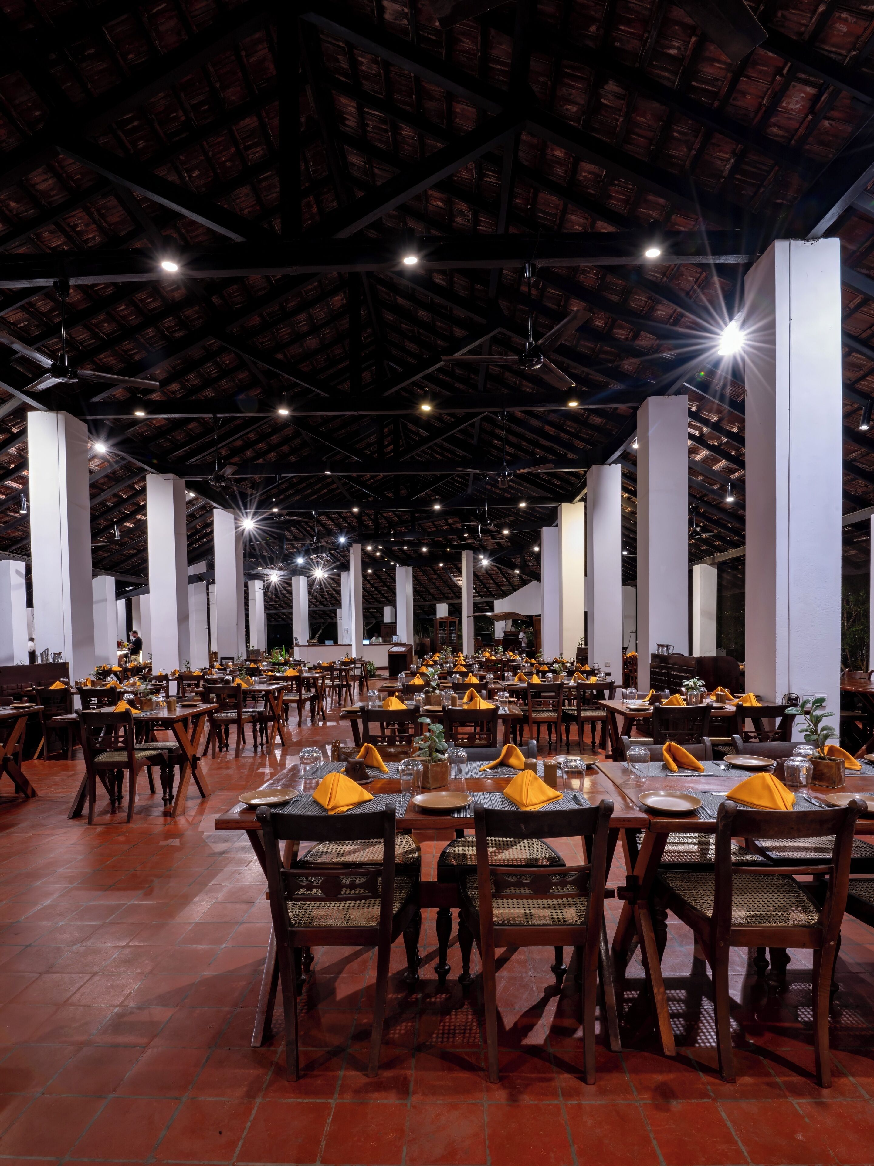 Un restaurant spacieux avec de hauts plafonds et des poutres apparentes, où de nombreuses tables sont dressées avec des serviettes jaunes et des nappes blanches.