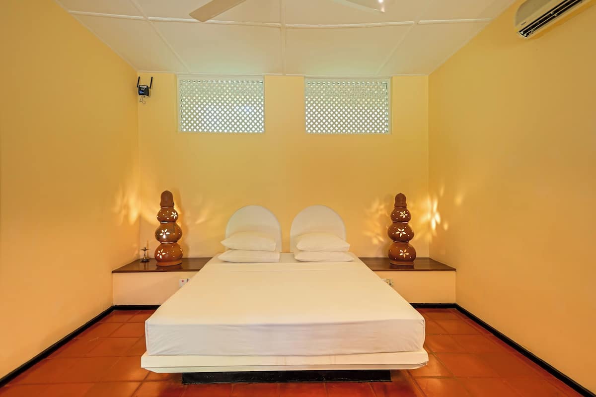 A neatly made bed in a room with two windows, a ceiling fan, and an air conditioning unit.
