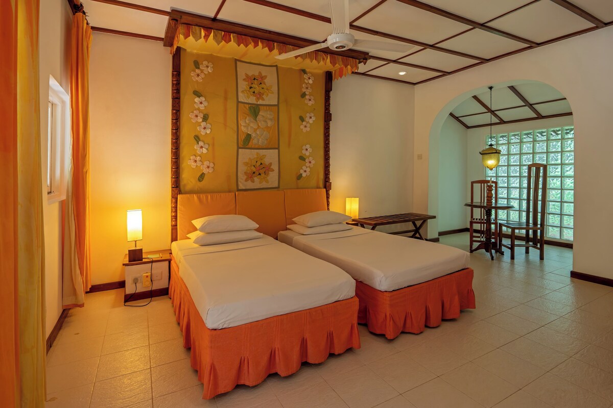 A hotel room with two beds, a ceiling fan, a table with chairs, and a view of greenery through a window.