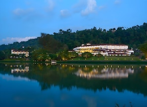 A large resort with multiple buildings, a lake in the foreground, and a forested hill in the background. - Cinnamon Citadel Kandy (Kandy)