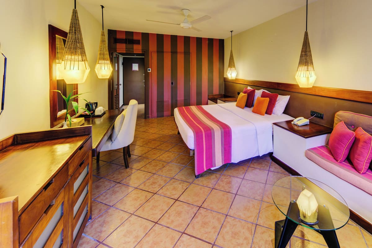 Une chambre d’hôtel avec un lit, un canapé, un bureau et une chaise. On y trouve un ventilateur de plafond et un luminaire fixé au mur.