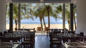 A restaurant with a view of the beach, large windows, and palm trees.