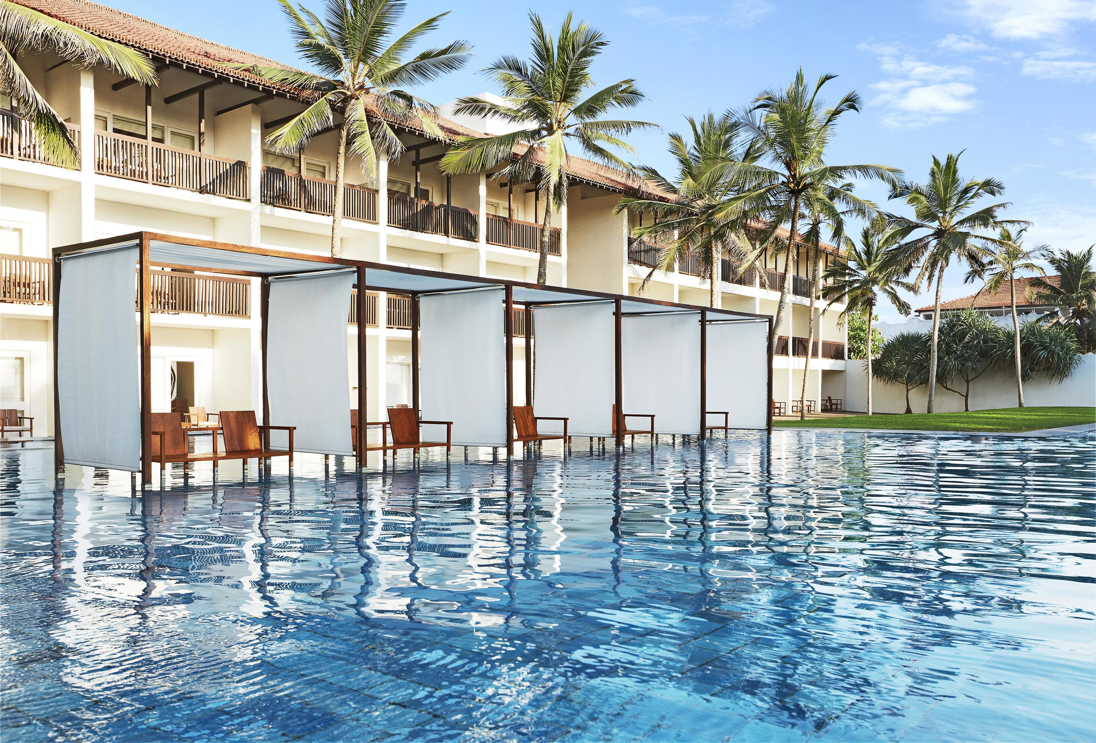 A swimming pool with lounge chairs and a covered seating area, surrounded by palm trees and a building with balconies.