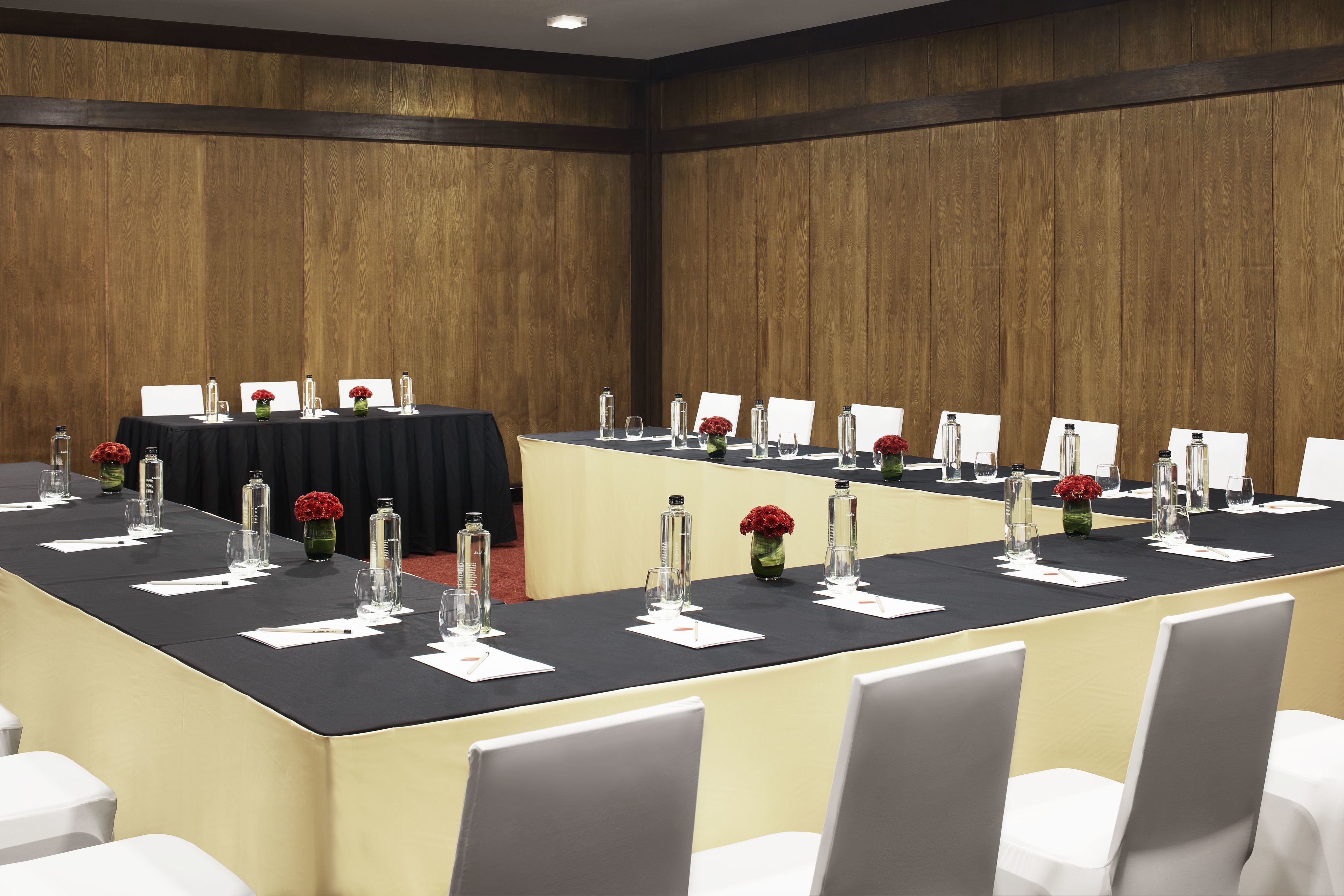 Un salón de conferencias con una larga mesa, sillas blancas y flores rojas en jarrones.