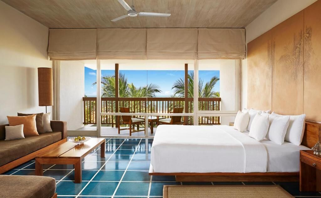 A hotel room with a bed, sofa, and a balcony with a view of palm trees.