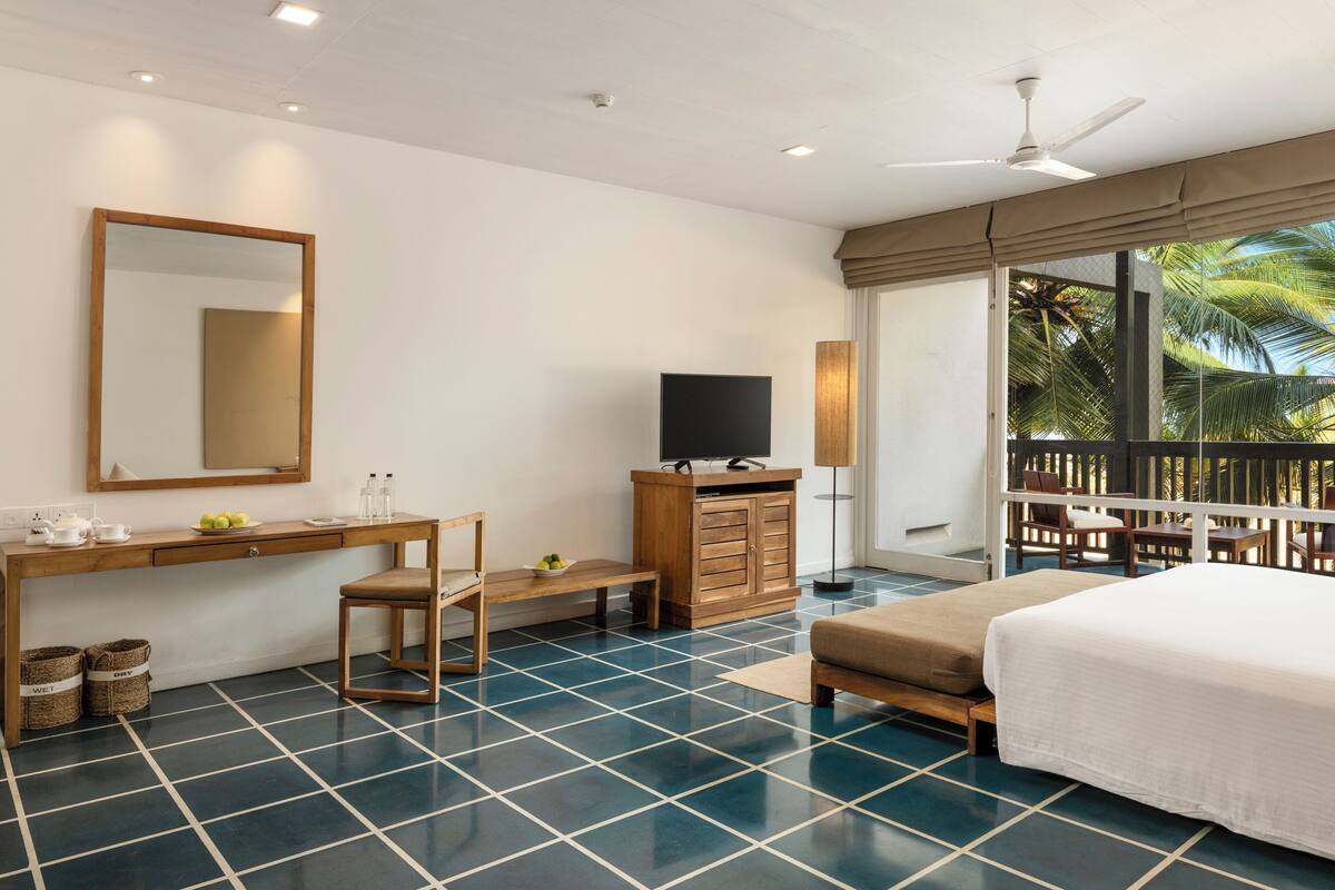 A hotel room with a bed, a desk with a television, a chair, a small table, and a balcony with a view of palm trees.