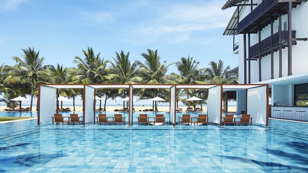 A swimming pool with lounge chairs and umbrellas, surrounded by palm trees and a building with balconies.