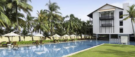 A swimming pool with lounge chairs and umbrellas, surrounded by palm trees and a modern building with balconies.