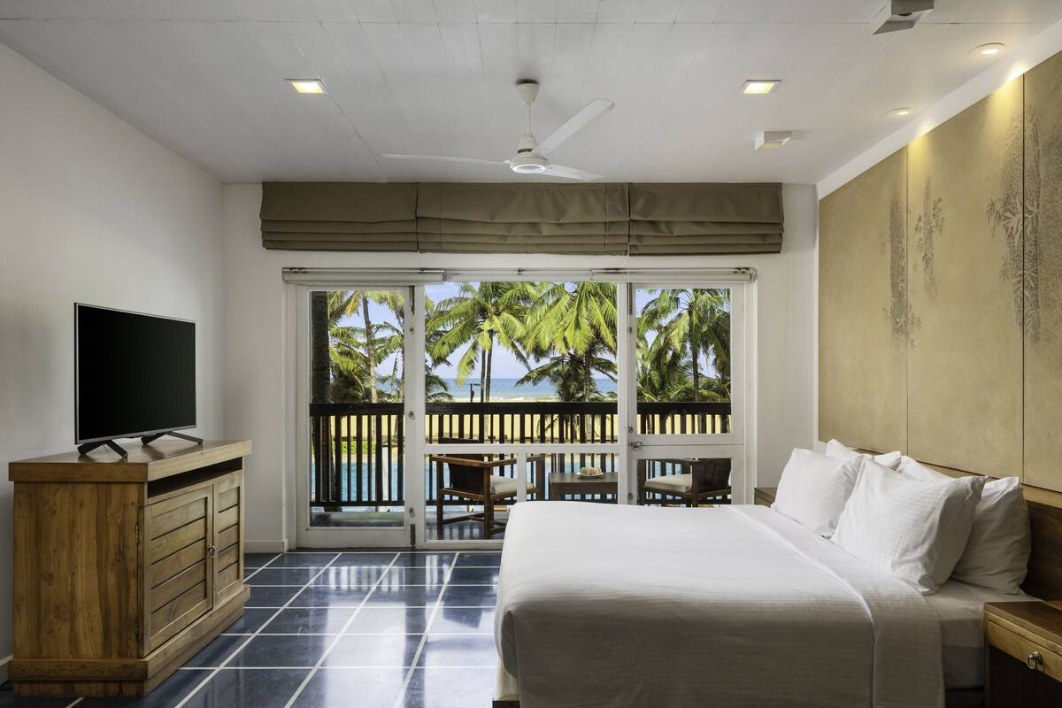 A bedroom with a bed, a TV, a wooden dresser, and a balcony with a view of palm trees and the ocean.