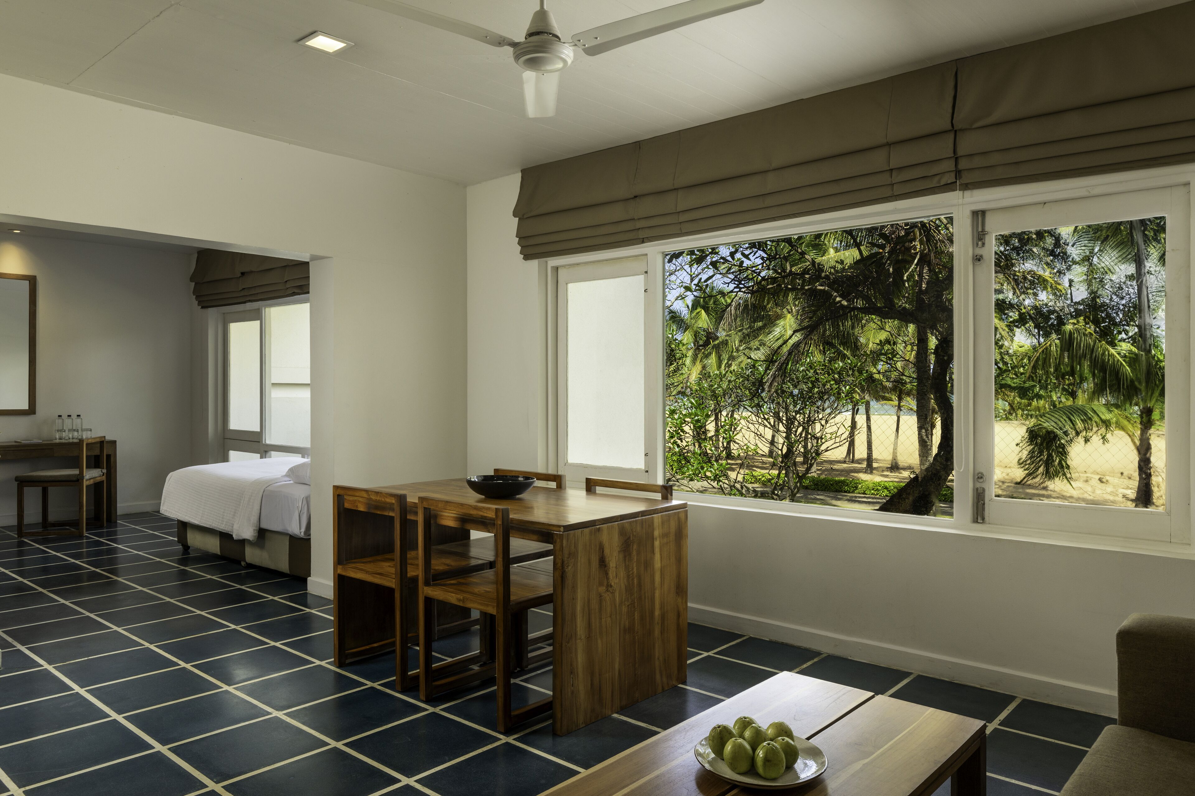 Una habitación con una cama, una mesa de comedor con sillas, una mesita con un frutero y una ventana que ofrece vistas a una playa y palmeras.