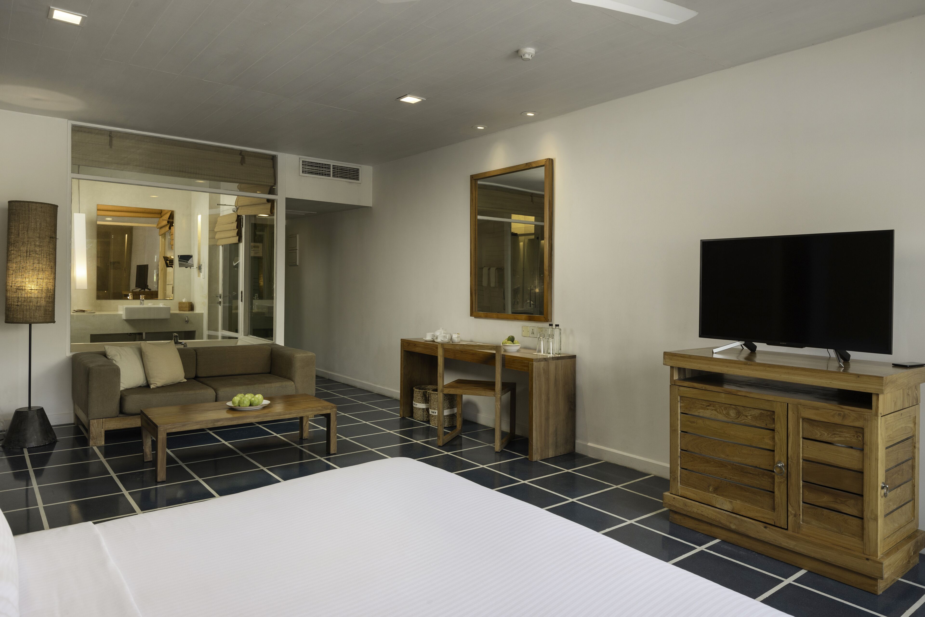 Une chambre d’hôtel moderne avec un grand lit, une commode en bois, une télévision, un canapé et une petite table sur laquelle se trouve un saladier rempli de fruits.