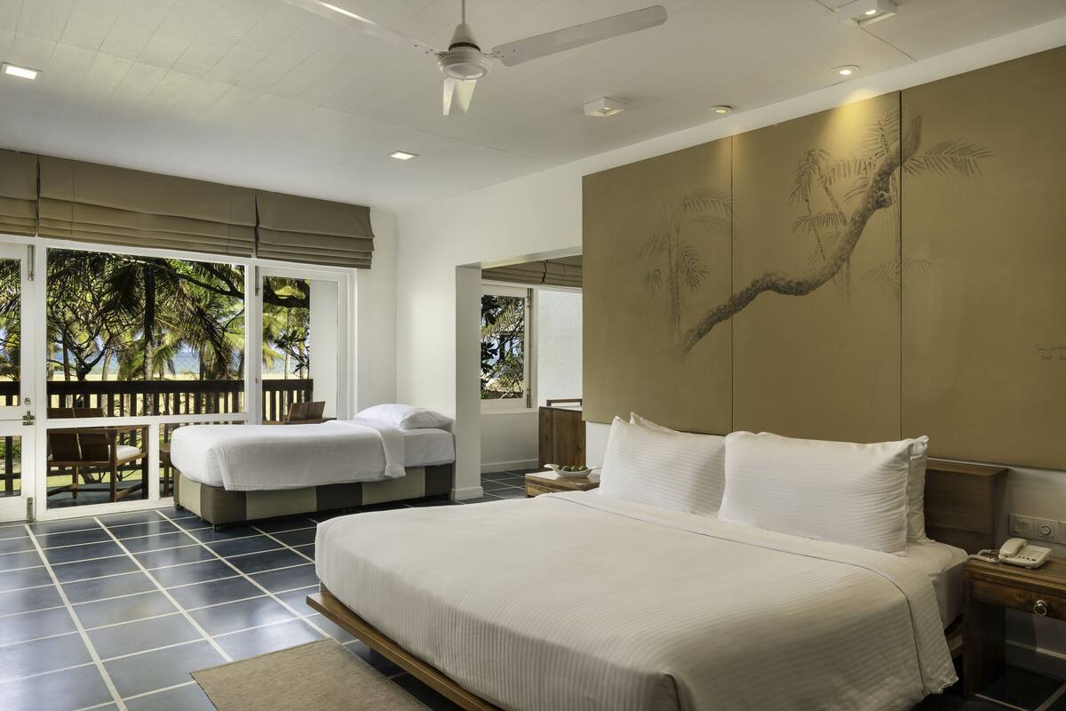 A hotel room with a large bed, a smaller bed, a desk, and a view of palm trees through the windows.