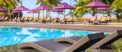 Un espace aménagé au bord de la piscine, avec des chaises longues et des parasols violets, le tout entouré de palmiers.