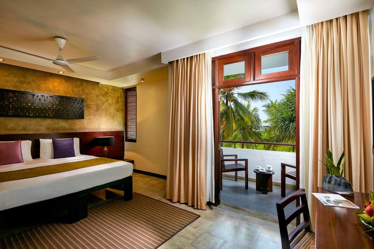 A hotel room with a bed, a ceiling fan, a view of palm trees, and a balcony with a table and chairs.