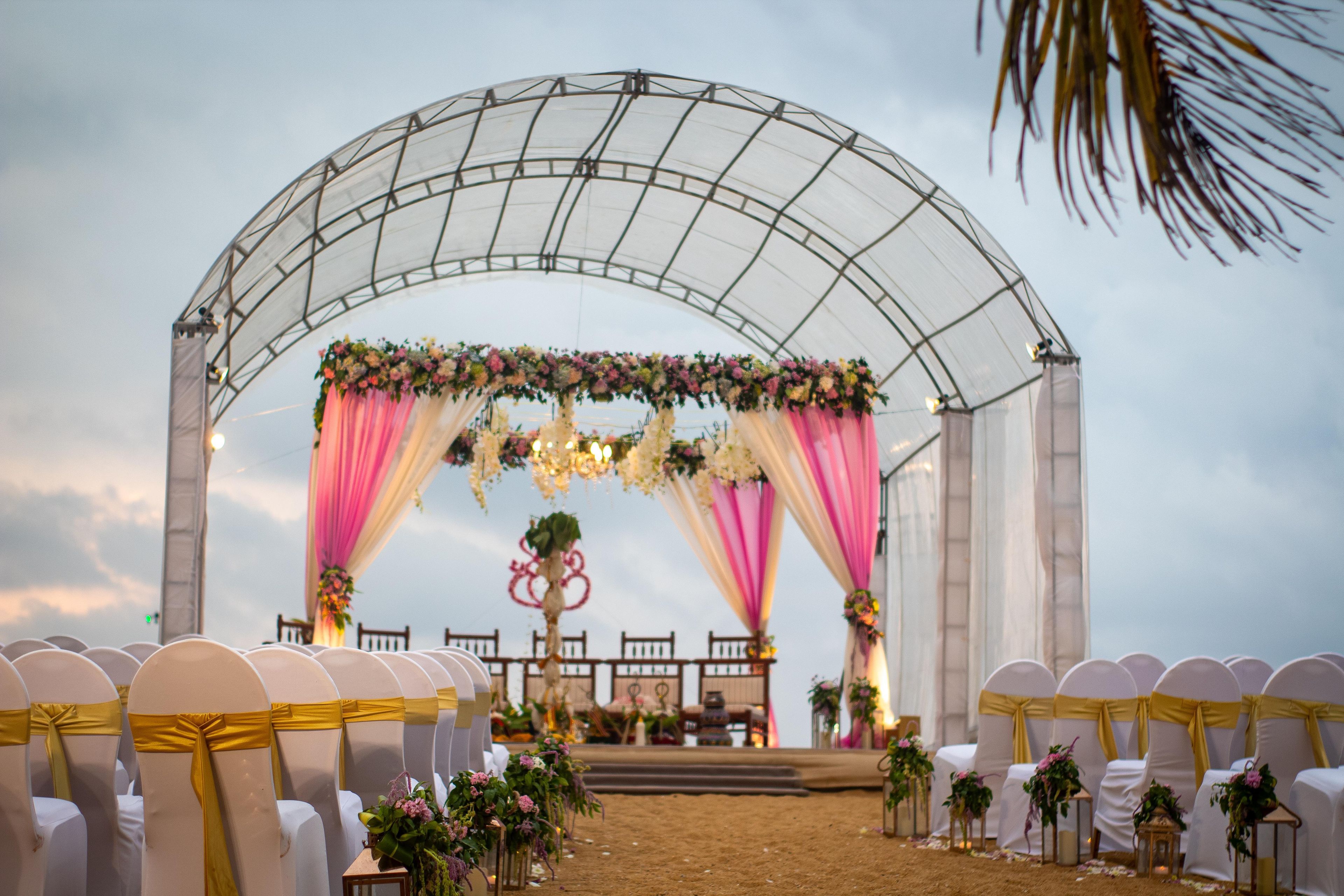 An outdoor wedding ceremony setup with a covered stage, floral decorations, and rows of chairs with white covers and gold sashes.