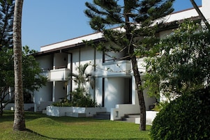 Um edifício moderno com telhado plano, paredes externas brancas e design simétrico. Possui várias varandas e um gramado bem cuidado com árvores.