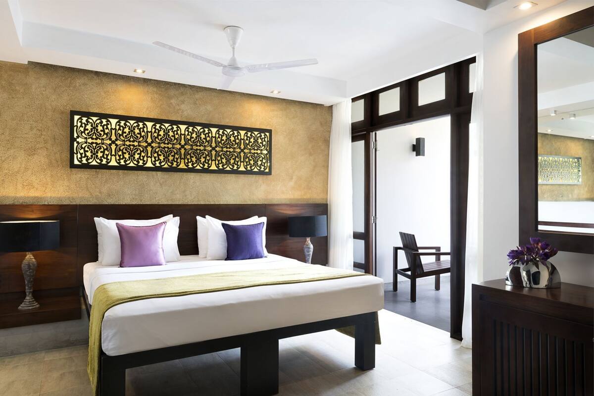 A modern hotel room with a large bed, a ceiling fan, a decorative wall piece, a desk with a vase, and a chair.