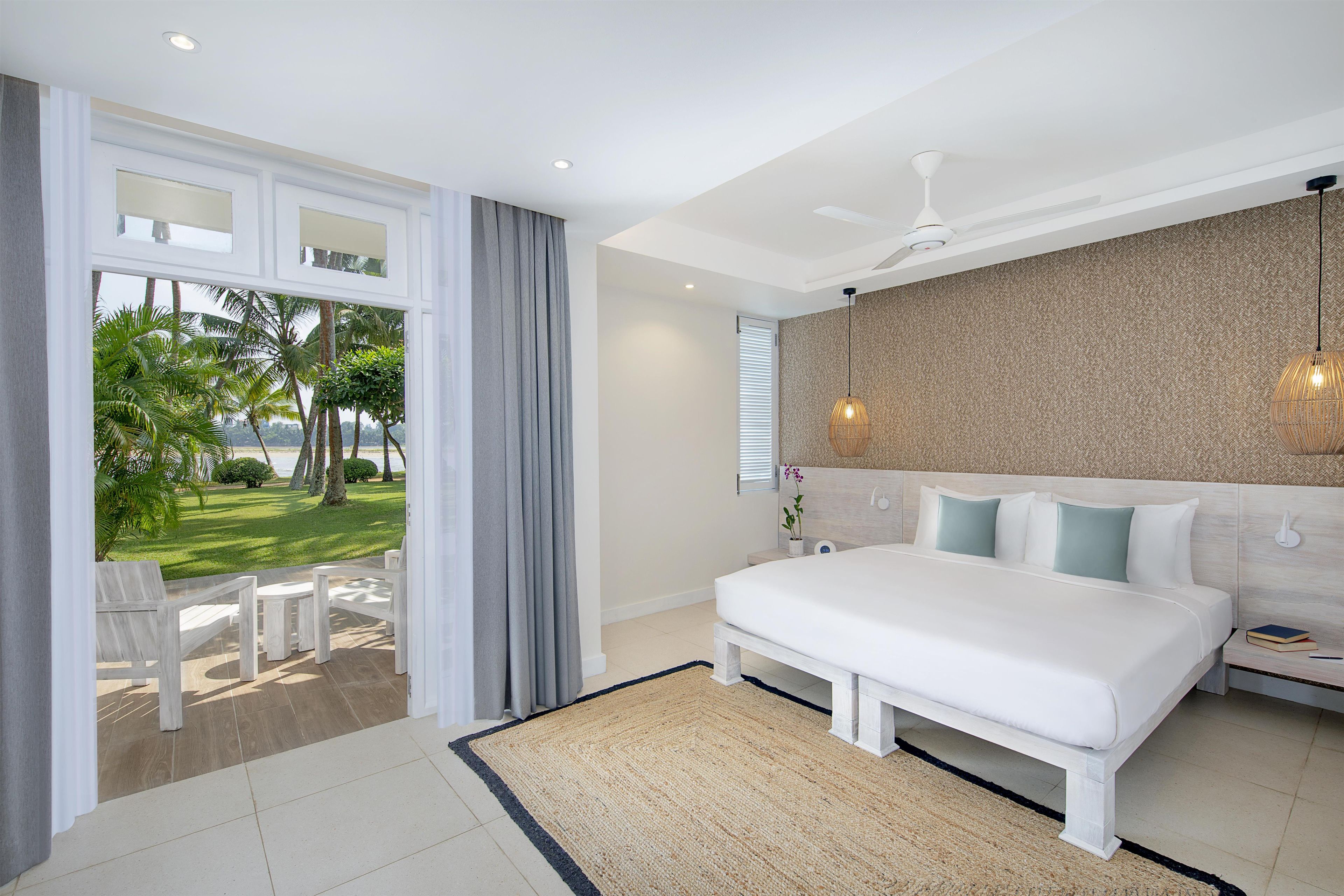 A bedroom with a large bed, a view of a garden, a ceiling fan, and a nightstand with a lamp.