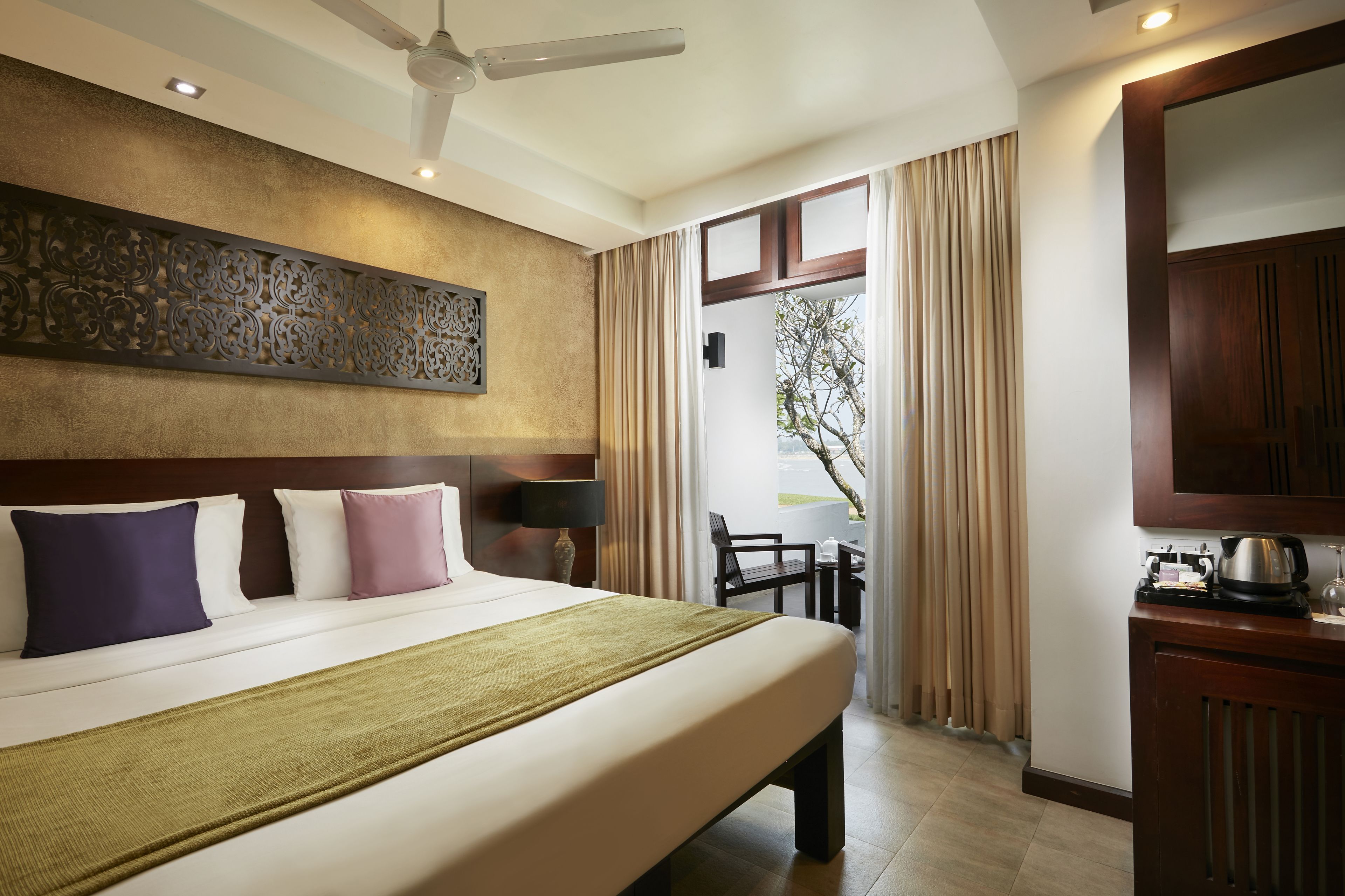 A hotel room with a large bed, a ceiling fan, a decorative wall panel, a view of the outdoors through a window, and a kitchenette area.
