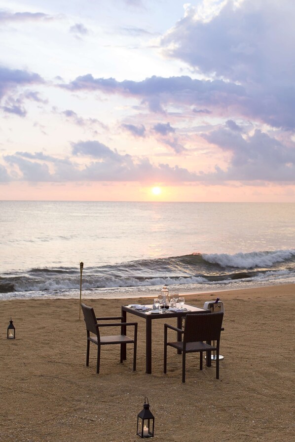 Une table dressée sur une plage de sable, avec des chaises, une lanterne et une vue sur l’océan et le coucher de soleil.