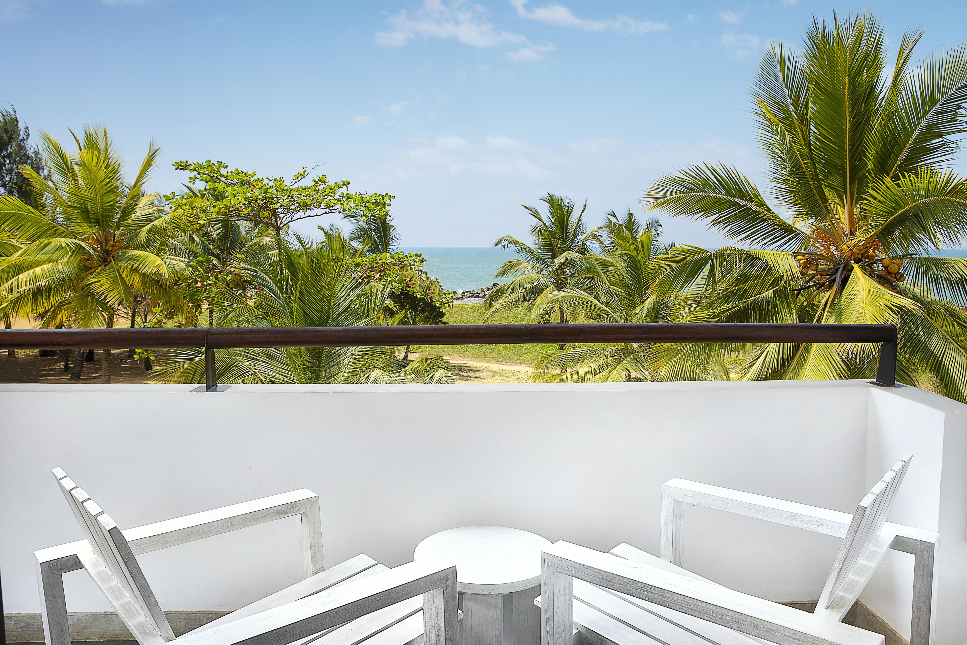 A balcony with a white table and chairs, overlooking a beach with palm trees and the ocean.