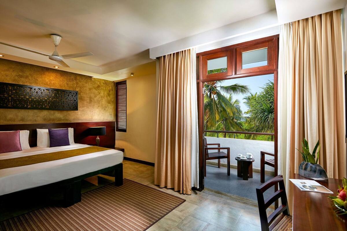 A hotel room with a bed, a ceiling fan, a window with curtains, a balcony with a table and chairs, and a decorative wall panel.
