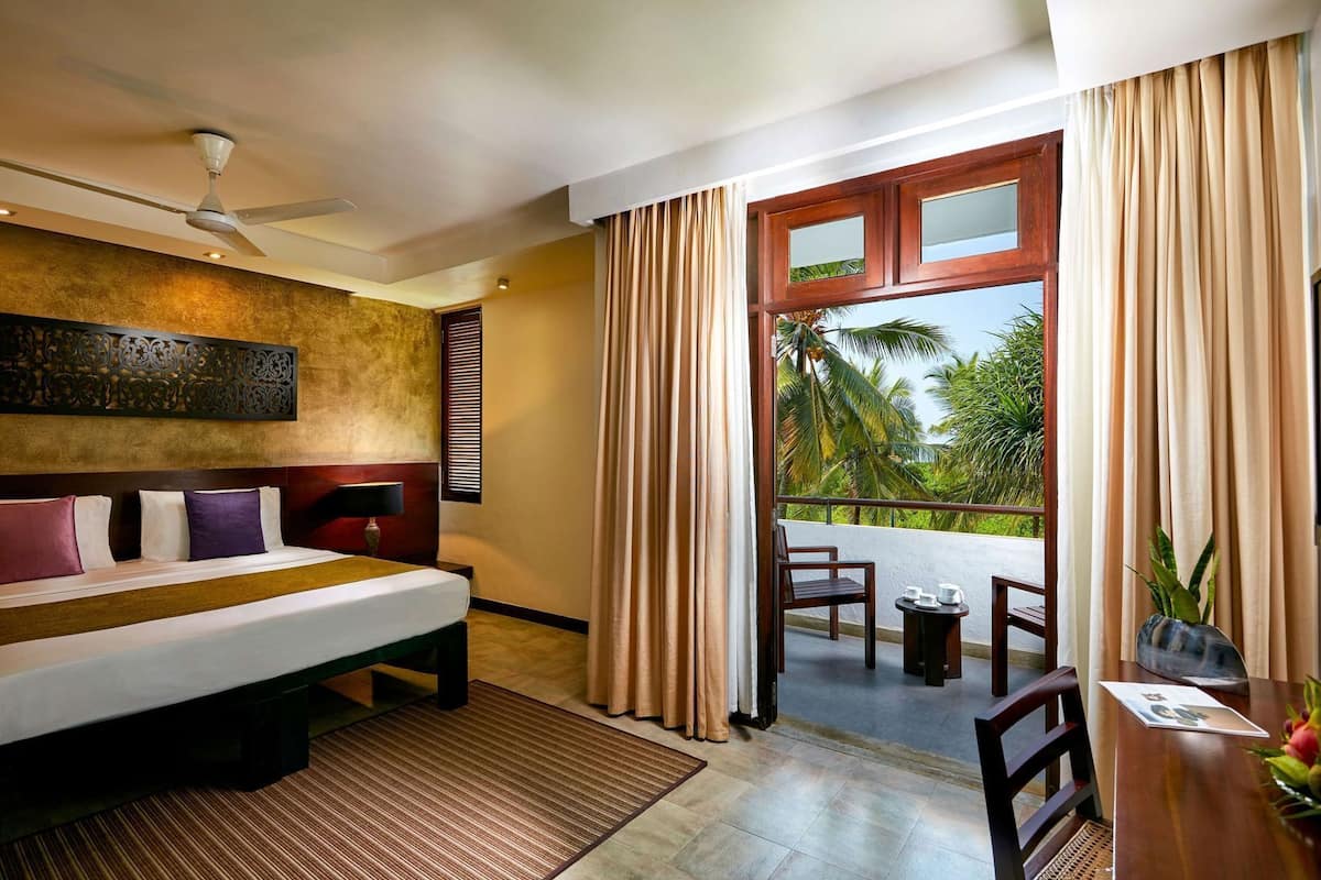 A hotel room with a bed, a ceiling fan, a window with curtains, a balcony with a table and chairs, and a decorative wall panel.