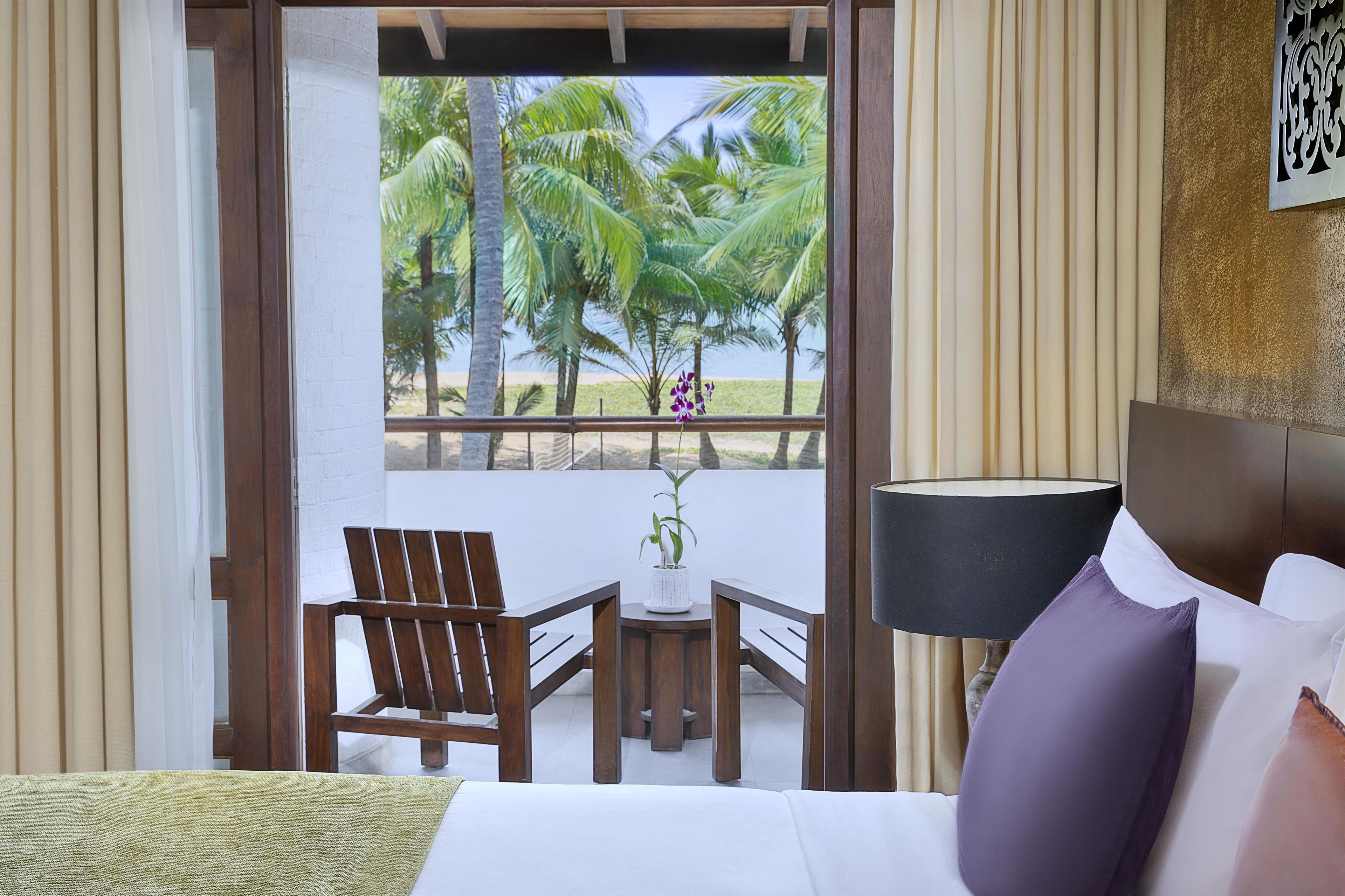 Habitación de hotel con una cama, una mesita con una planta, una silla y vistas a palmeras y a la playa.