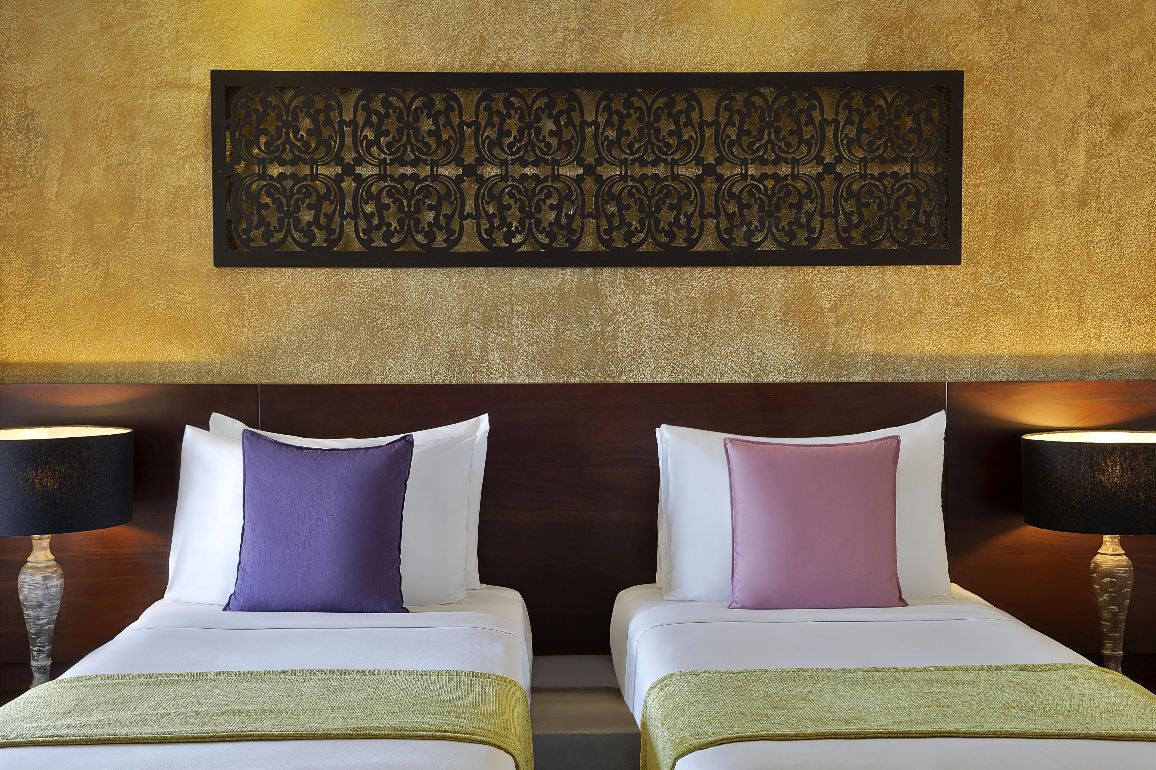Habitación de hotel con dos camas, cada una con ropa de cama blanca y una almohada morada. Hay una pieza decorativa en la pared sobre las camas y una lámpara a cada lado.