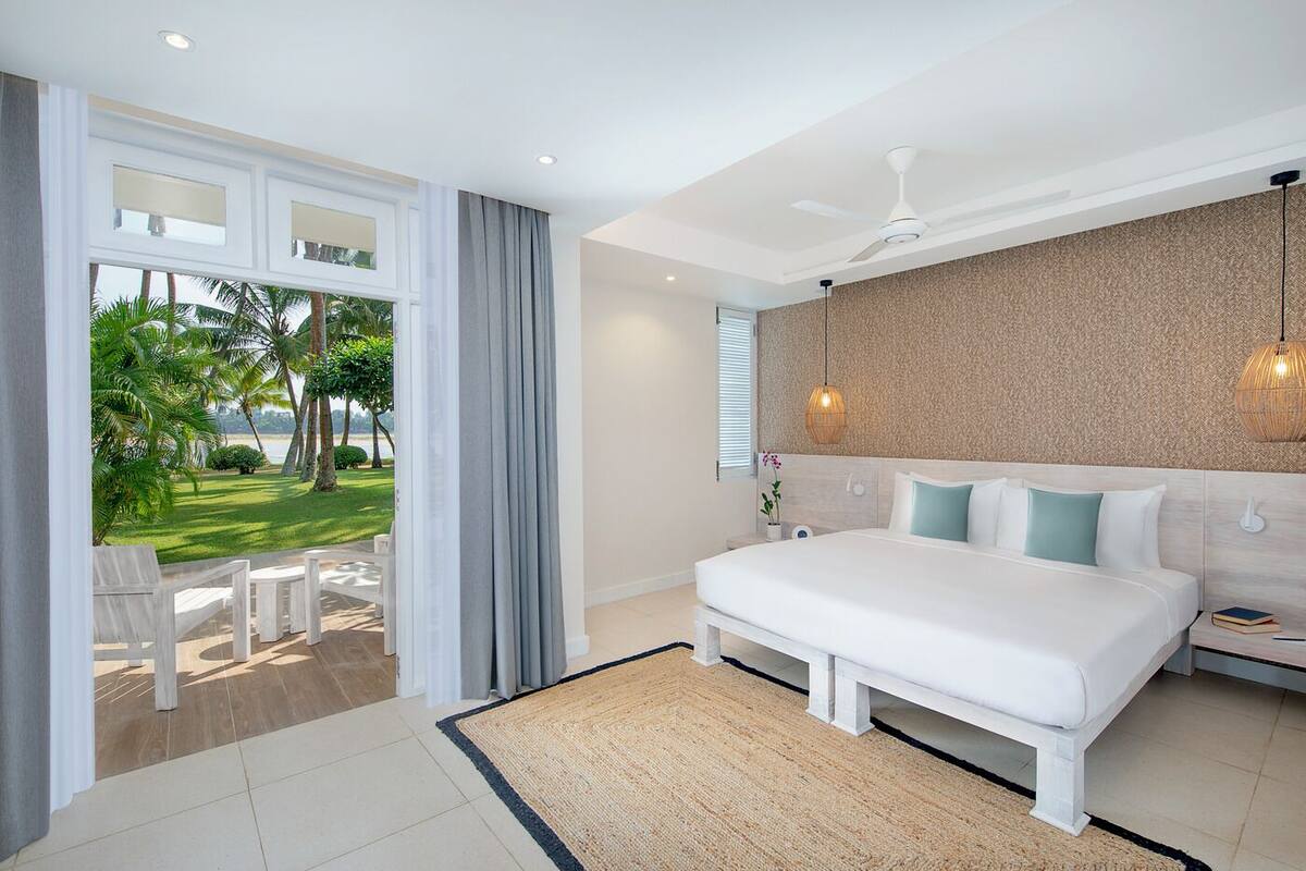 A bedroom with a large bed, a ceiling fan, and a view of a garden with palm trees.