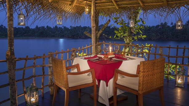 A dining area with a table set for two, surrounded by wooden chairs, under a thatched roof with hanging lanterns. - Avani Kalutara Resort (Kalutara)