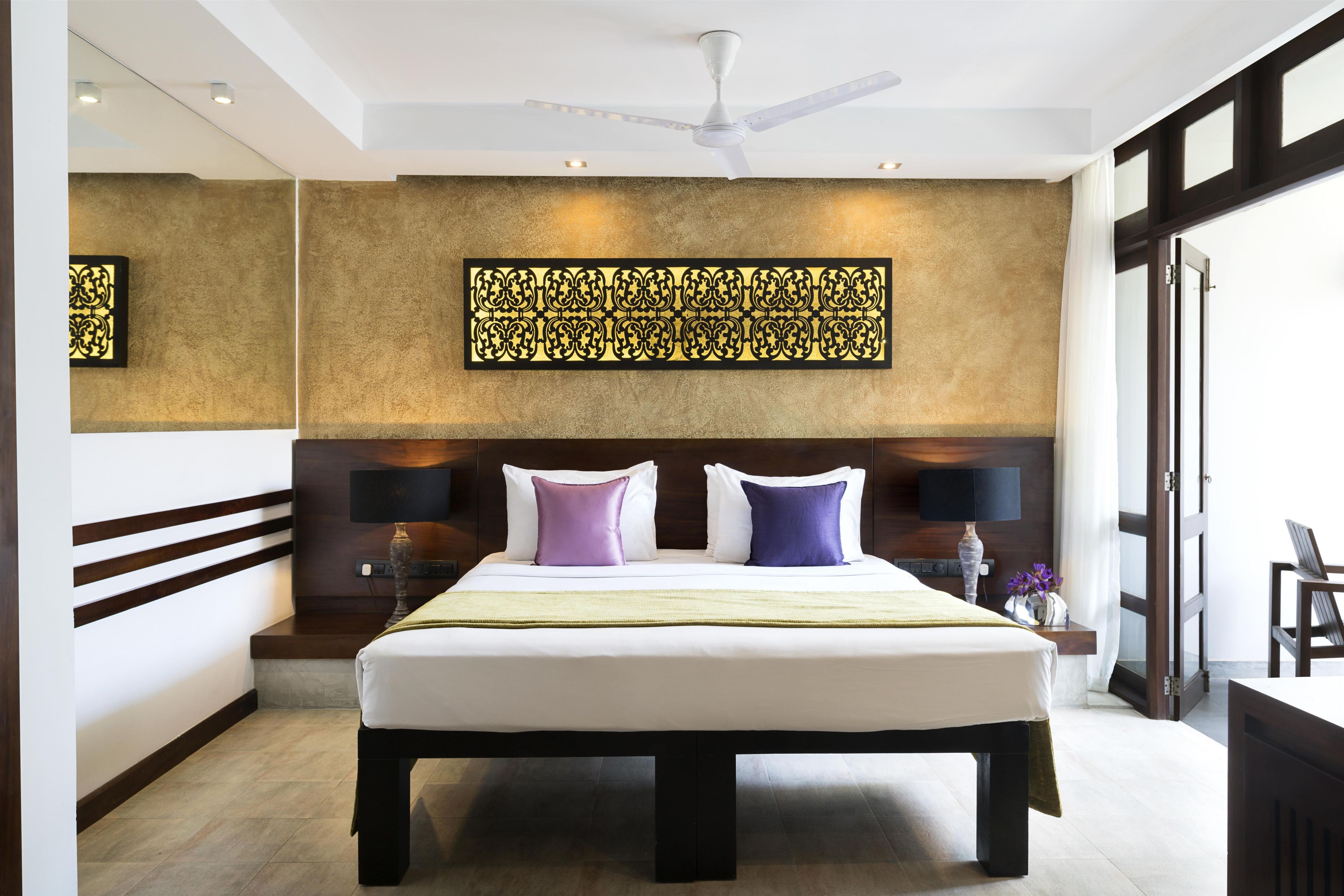A hotel room with a large bed, a ceiling fan, a decorative wall panel, and a bedside table with a lamp.