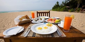 A breakfast setup on a wooden table with plates of food, including eggs, shrimp, and toast, and a glass of orange juice on a beach. - Nilaveli Beach (Nilaveli)