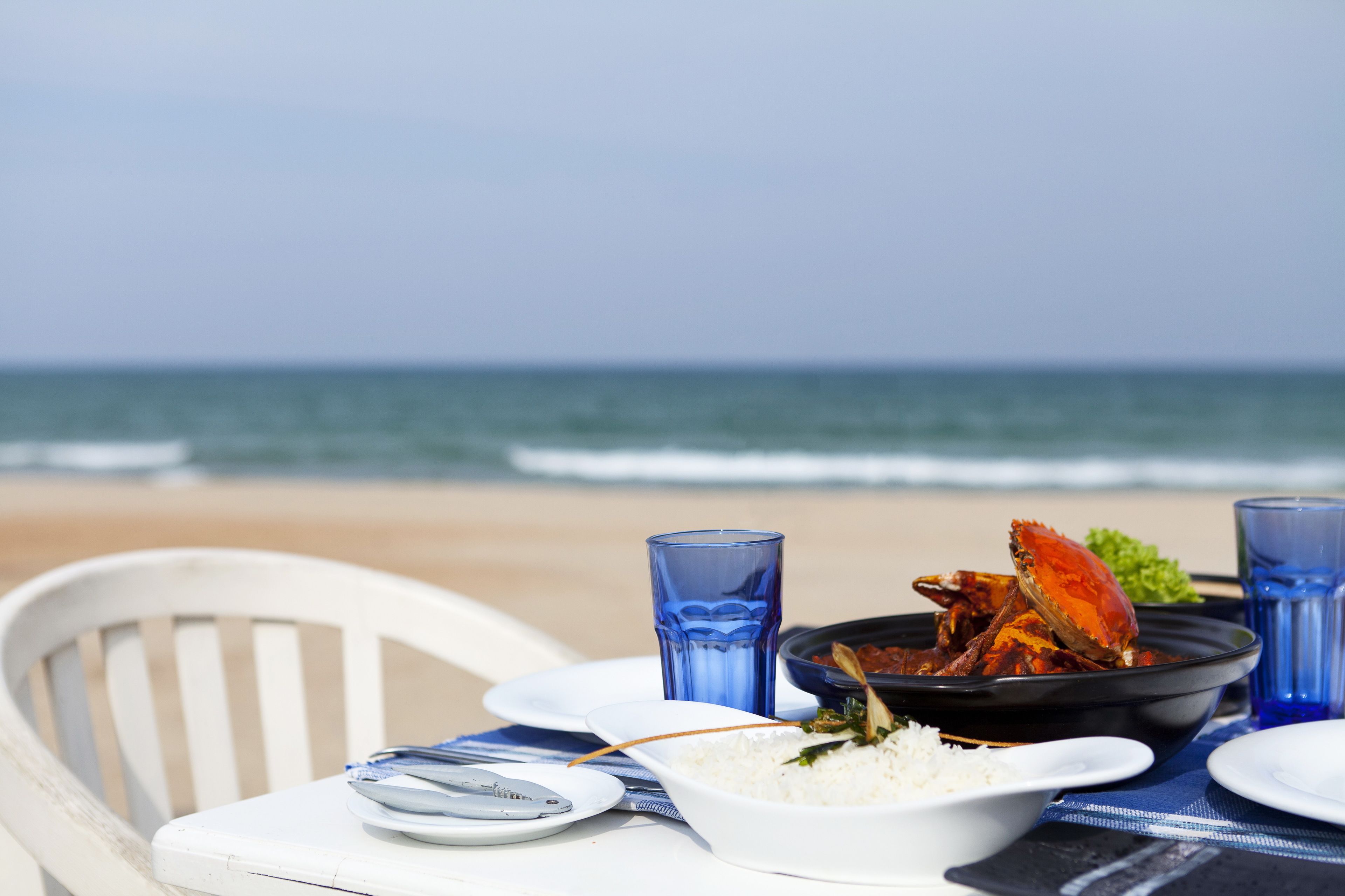 Mesa con un tazón de arroz, un plato de cangrejo y vasos azules en una playa, con una silla.