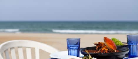 Mesa con un tazón de arroz, un plato de cangrejo y vasos azules en una playa, con una silla.