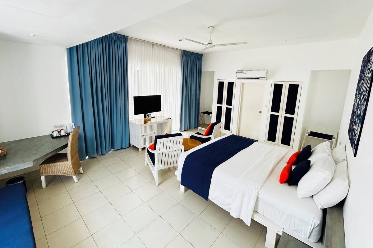 A hotel room with a bed, a desk with a television, a chair, a wardrobe, and a ceiling fan.