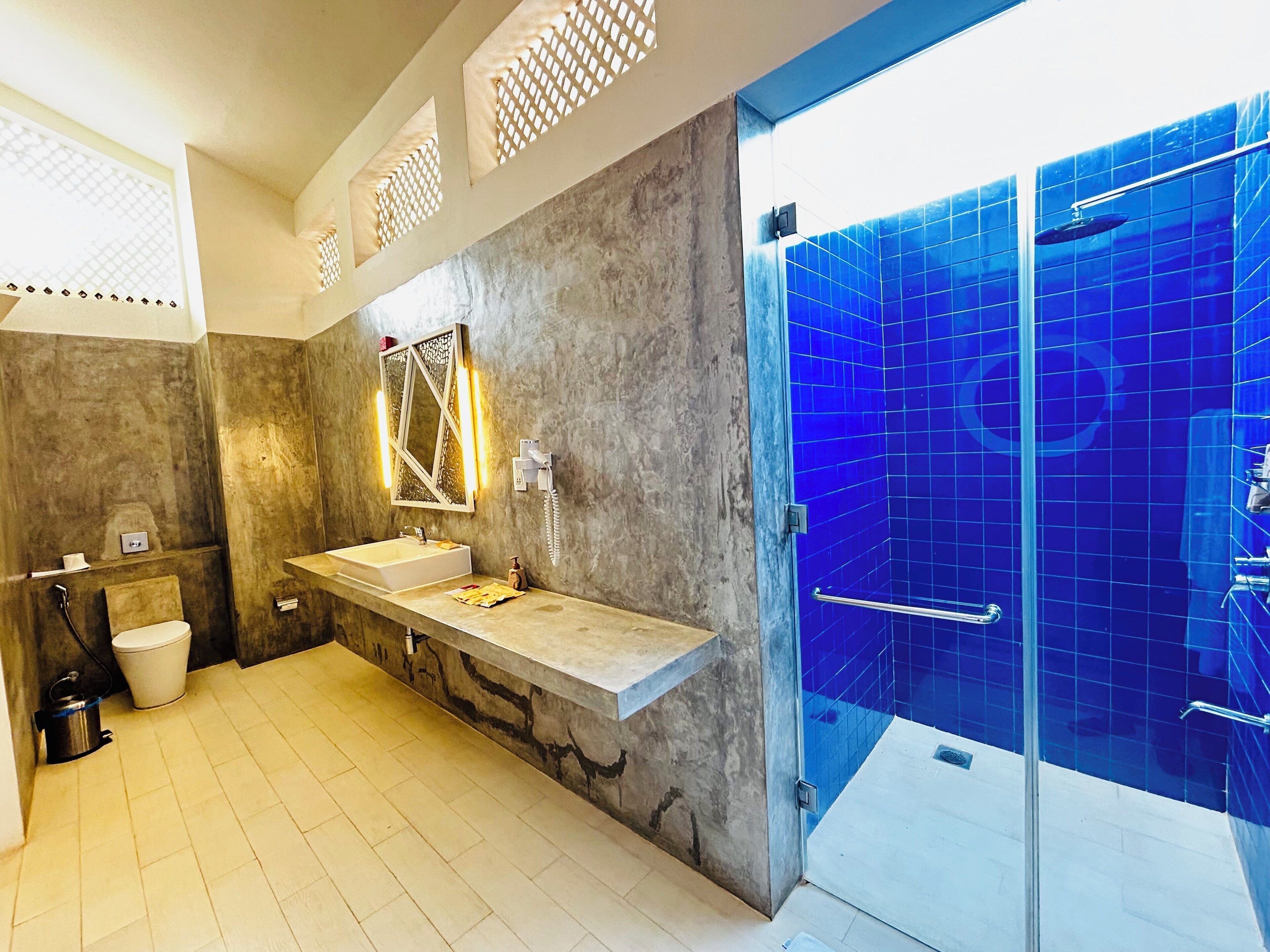 Ein modernes Badezimmer mit einem großen Spiegel, zwei Waschbecken, einer Toilette und einer begehbaren Dusche mit blauen Fliesen.